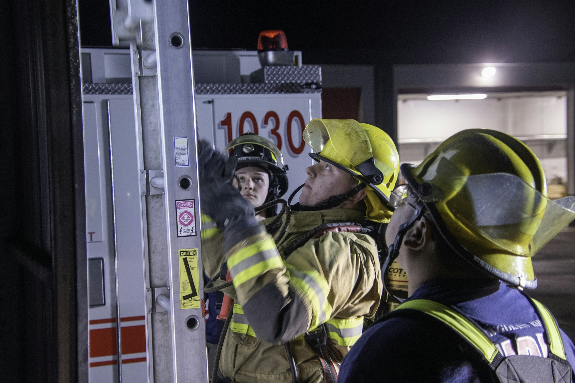 Three firefighters in helmets and gear focus on a ladder at night, with an emergency vehicle visible in the background.