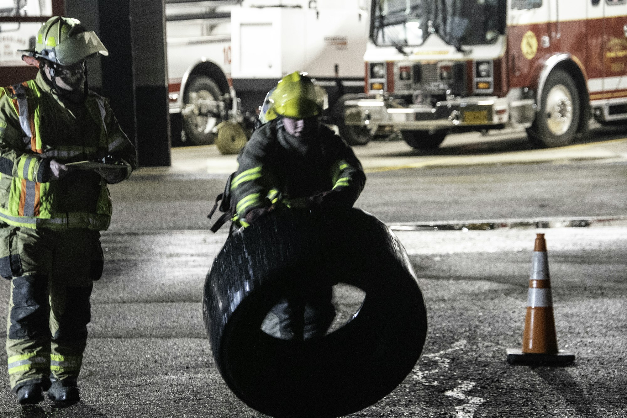 Two firefighters are working at a station; one is carrying a tire while the other holds a clipboard. A fire truck is in the background.