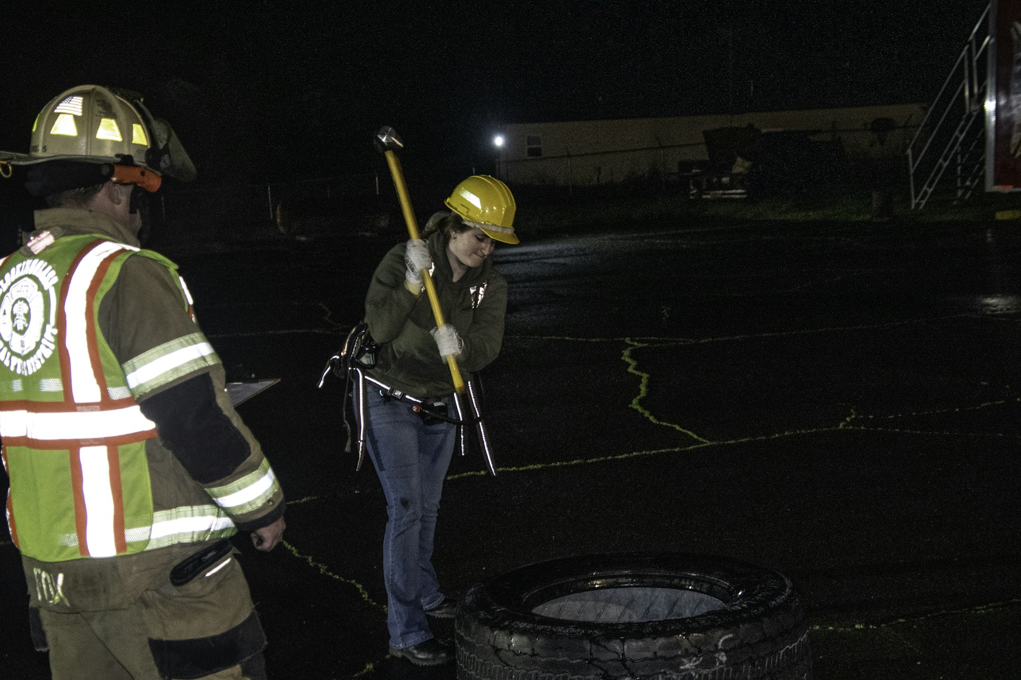 A person in a hard hat swings a tool towards a tire, while another in firefighter gear observes in a darkened area.