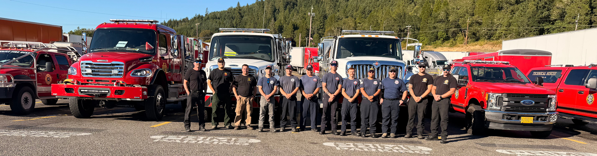 Firefighters stand in front of various fire trucks in a parking lot, showcasing their teamwork and equipment.
