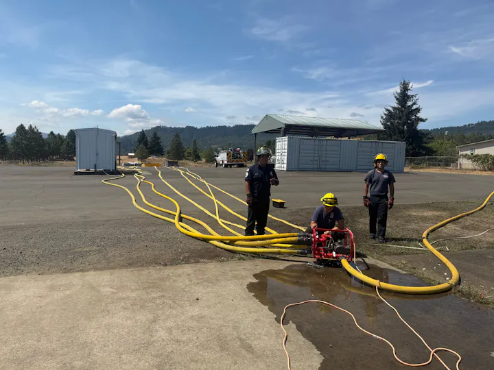 Firefighters are setting up equipment with yellow hoses and a pump at an outdoor training site under a clear blue sky.