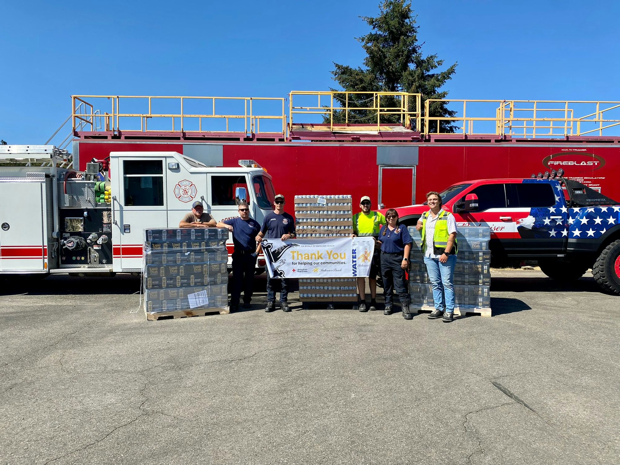Firefighters and volunteers with water packs and a "Thank You" banner, in front of a fire engine and decorated truck.