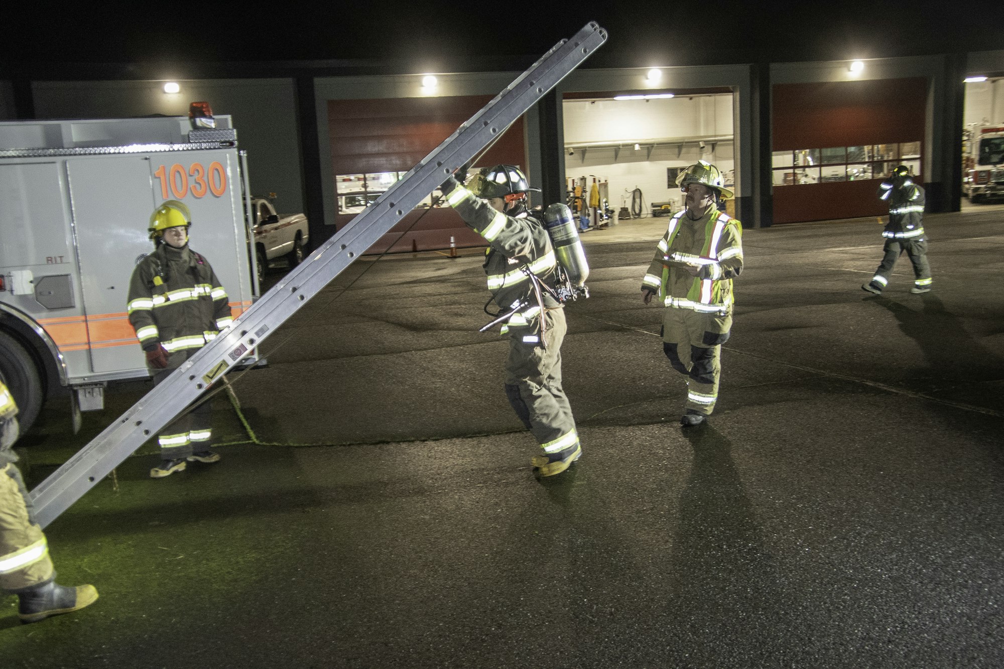 Firefighters are at night, setting up equipment, including a ladder, near fire trucks. Safety gear is visible on all personnel.