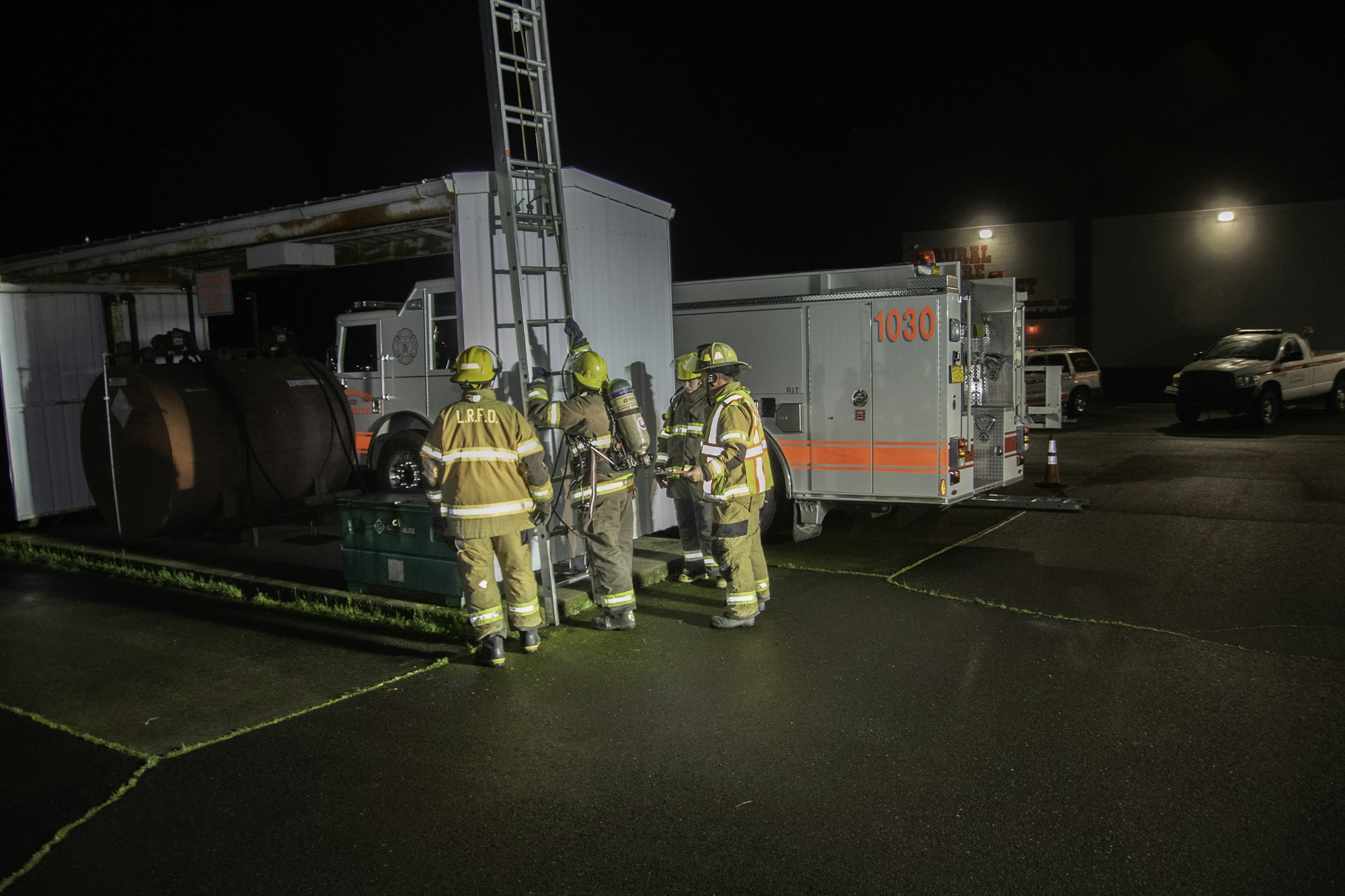 Firefighters at night are responding to a situation, equipped with gear and a ladder, near emergency vehicles and storage tanks.