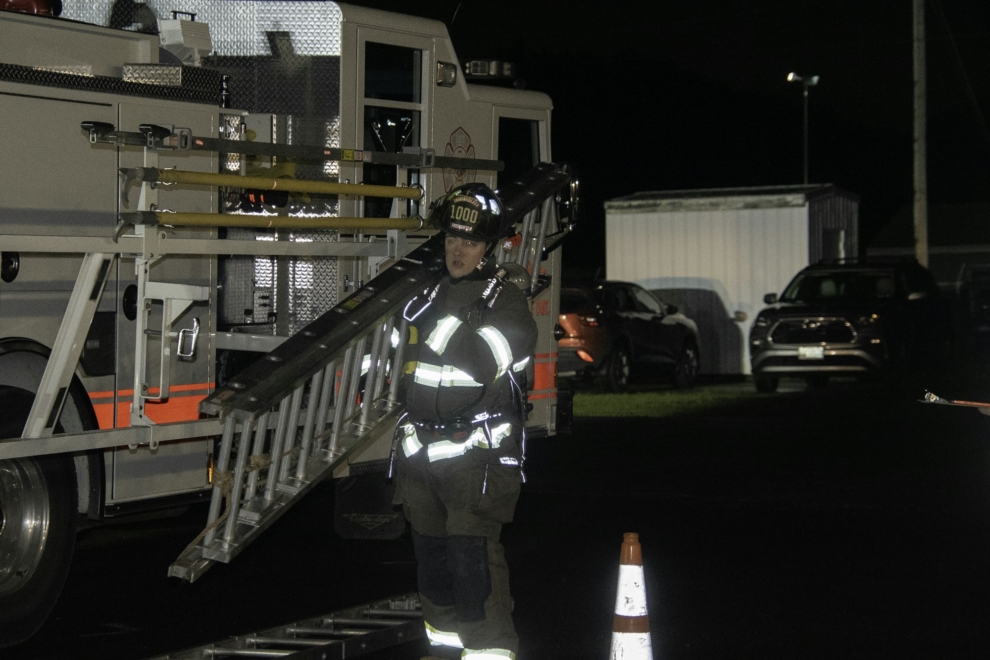 A firefighter carrying a ladder near a fire truck at night, with vehicles and a traffic cone also visible in the background.