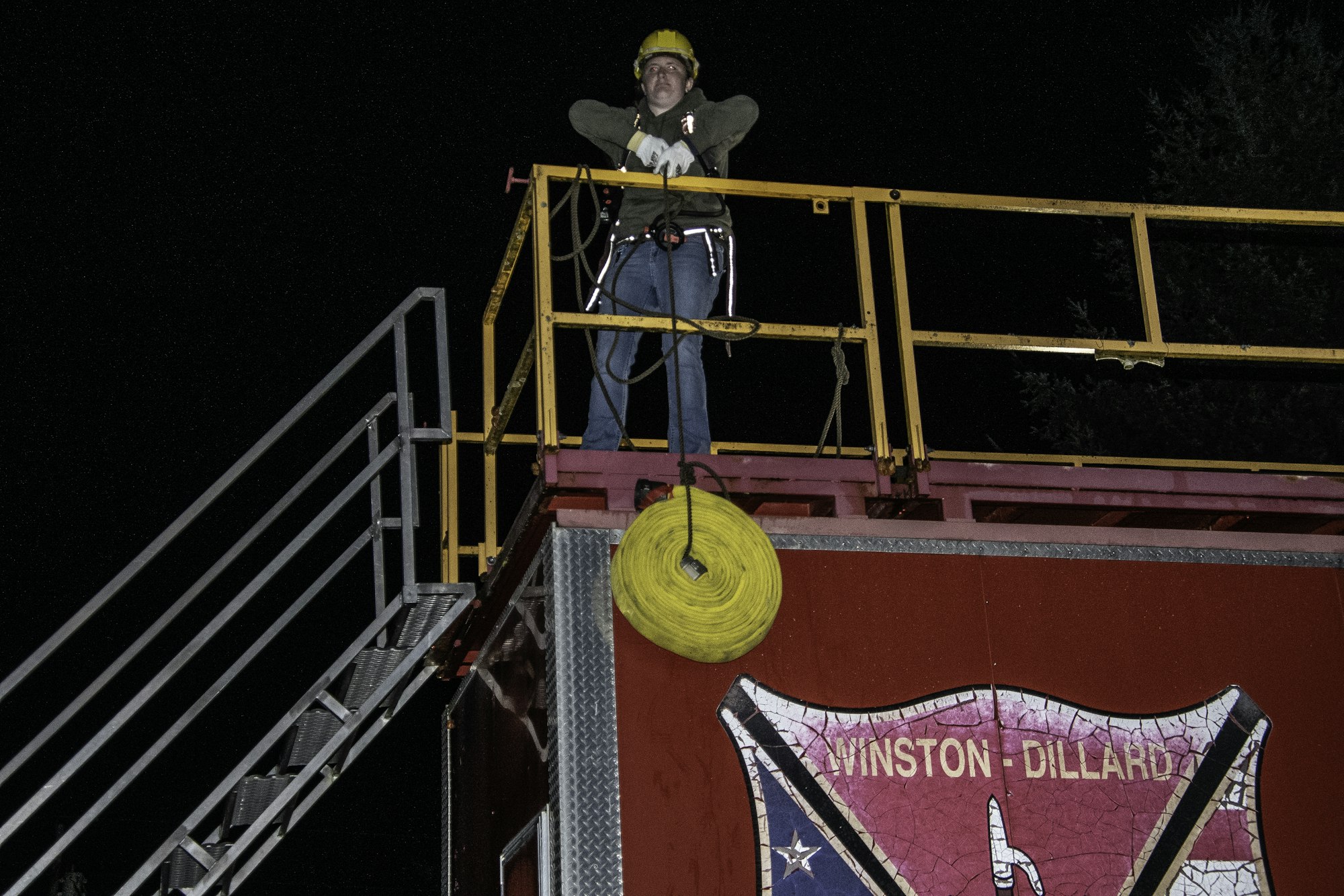A person in safety gear stands on a platform, overlooking the area, with equipment and tools around them at night.