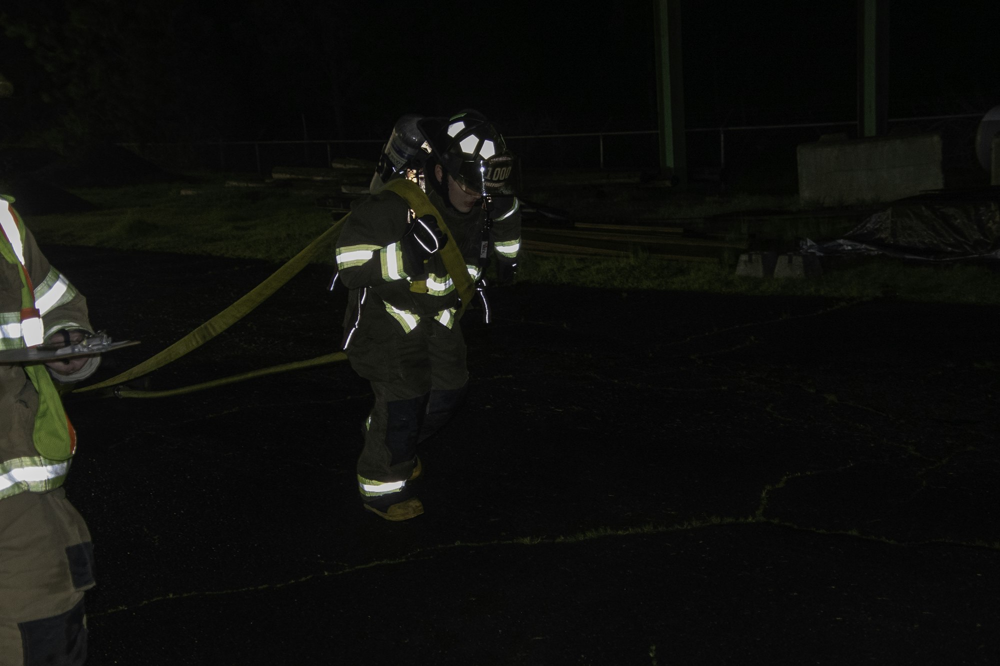 A firefighter in gear is carrying a hose at night, while another person observes with a clipboard nearby.