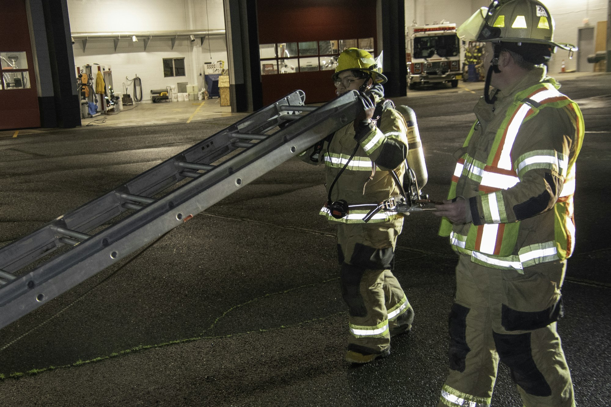 Two firefighters in gear are carrying a ladder outside of a fire station, preparing for an operation.