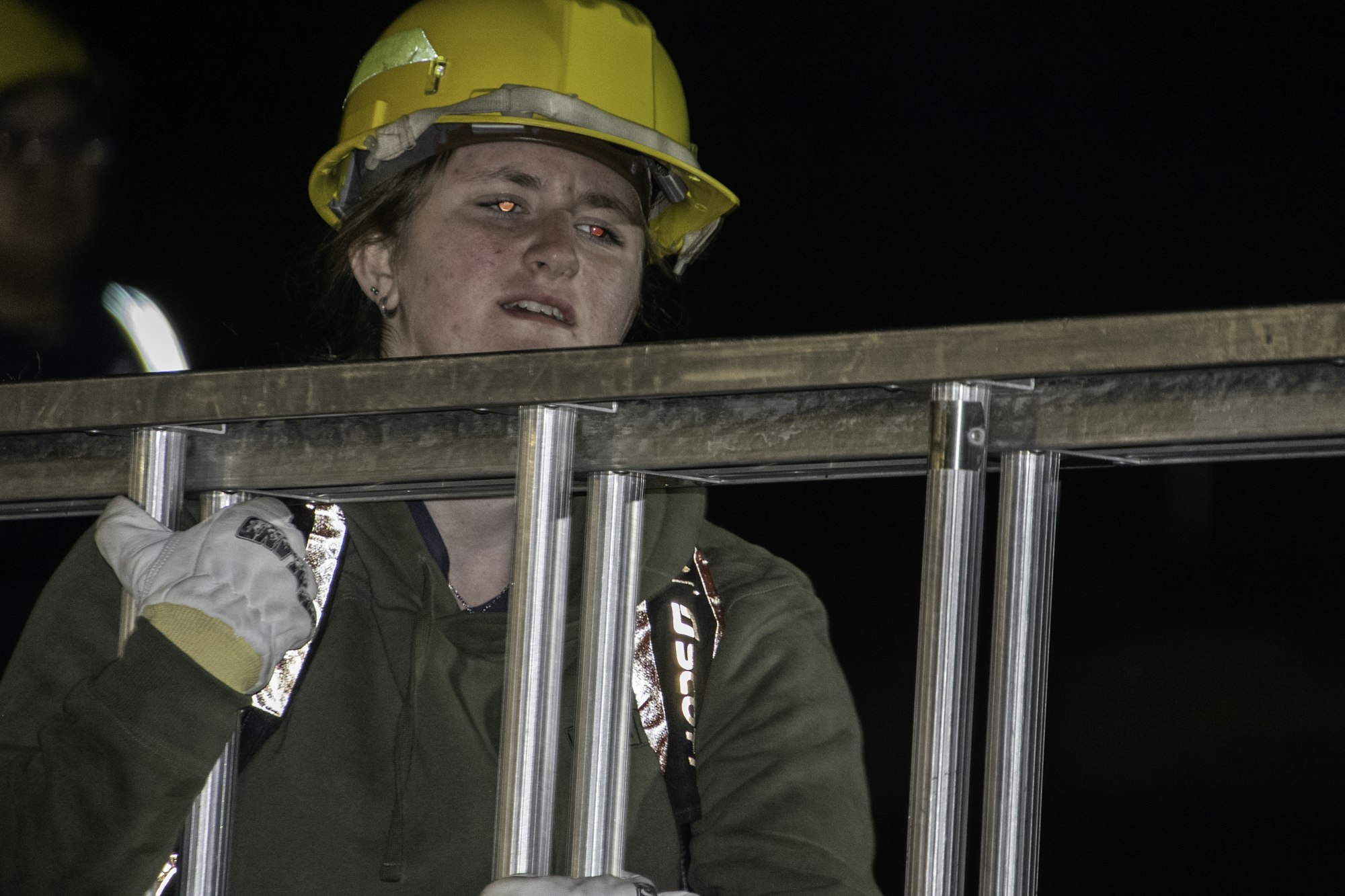 A person in a yellow hard hat is holding a metal ladder, wearing gloves, in a dark setting.