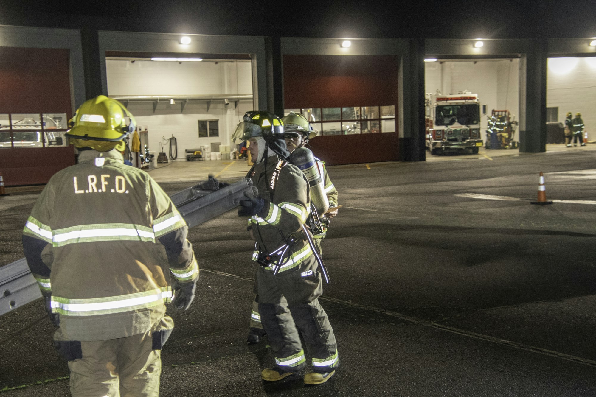 Firefighters in gear are working together, handling equipment outside a fire station at night.