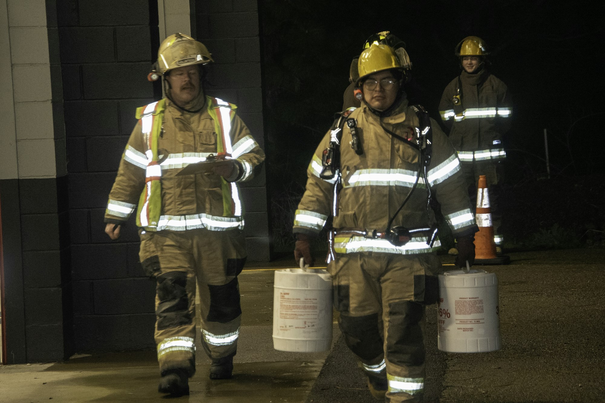 Two firefighters in gear walk with buckets, while another stands nearby, illuminated by nighttime lights.