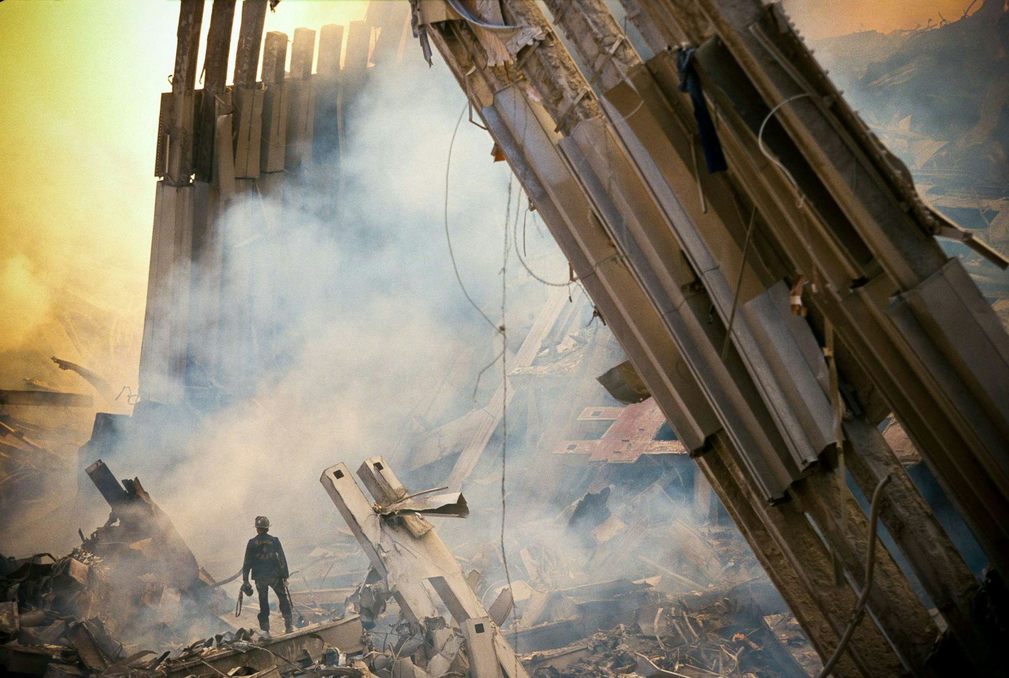 A lone person stands amidst smoky ruins and collapsed structures.