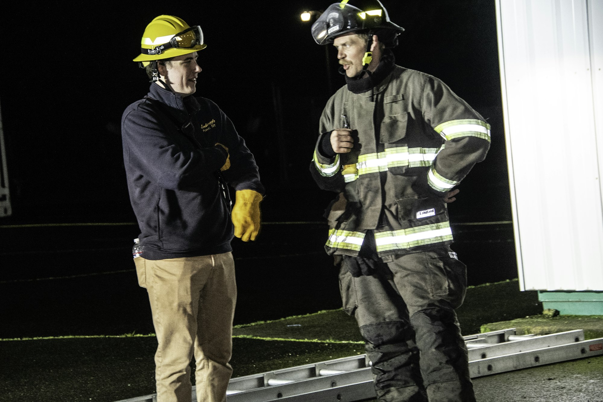 Two individuals are talking at night; one wears a firefighter's uniform and helmet, while the other is casually dressed.