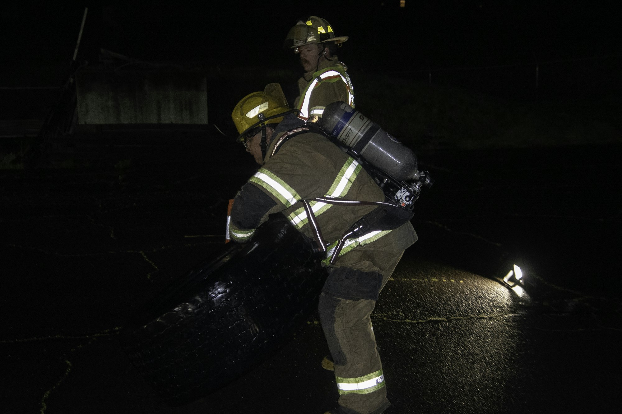 Two firefighters in protective gear are working together to lift a large tire at night, with lighting illuminating the scene.