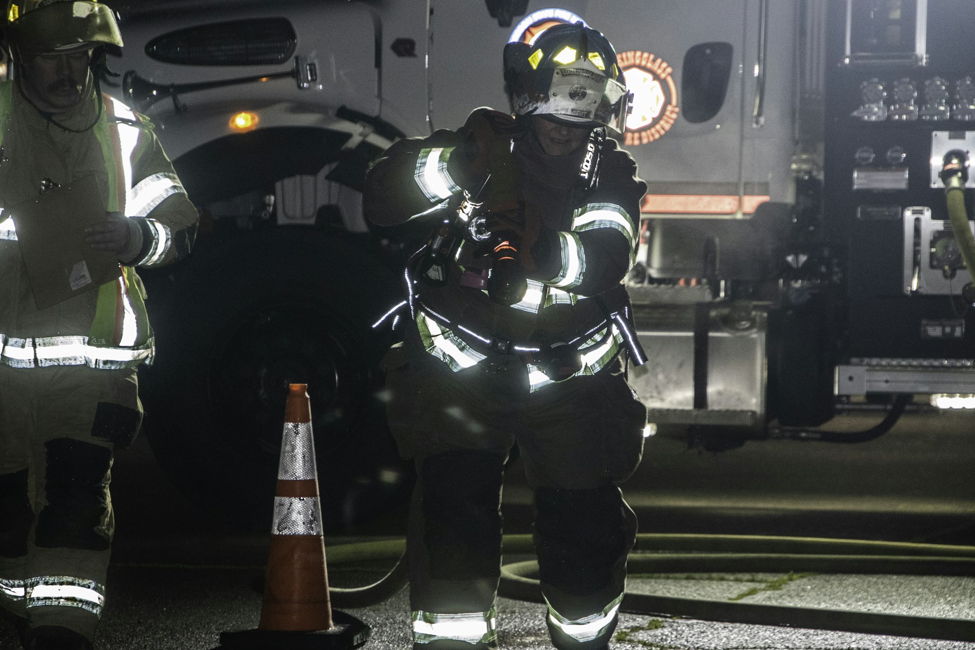A firefighter prepares equipment near a fire truck, with another firefighter nearby holding a clipboard, in a nighttime setting.