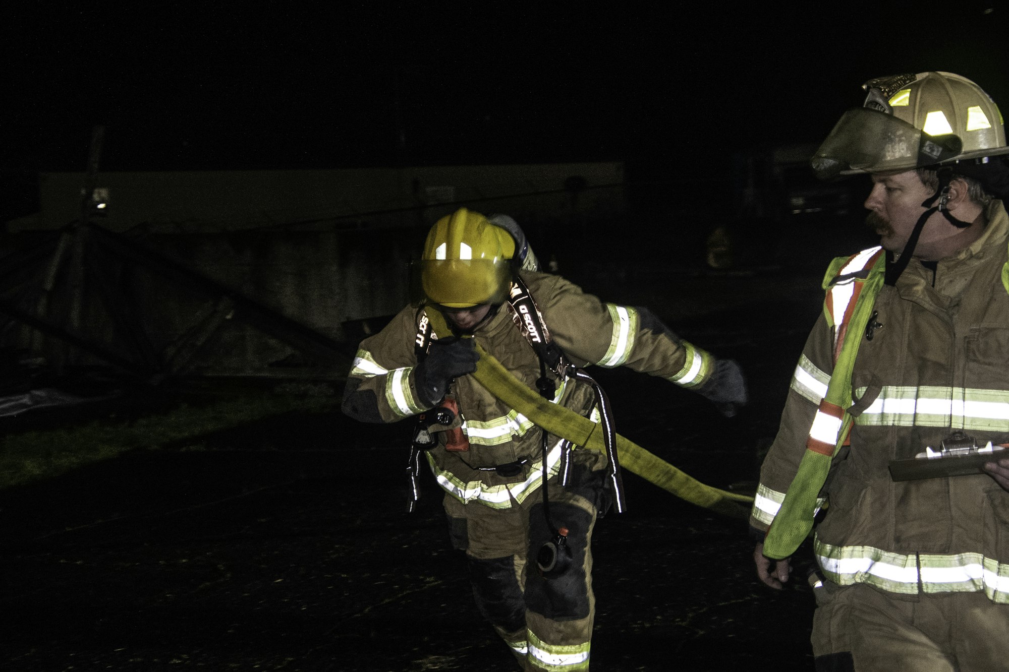 Two firefighters, one with a hose and one holding a clipboard, are engaged in a nighttime operation.