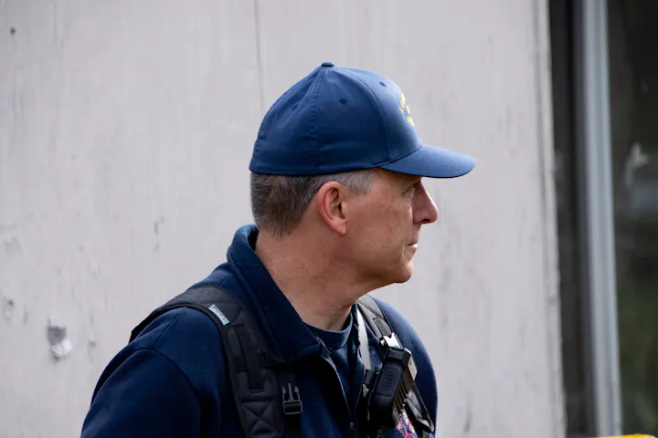 A man wearing a blue cap and dark jacket is seen in profile, with a radio on his shoulder, standing near a building.