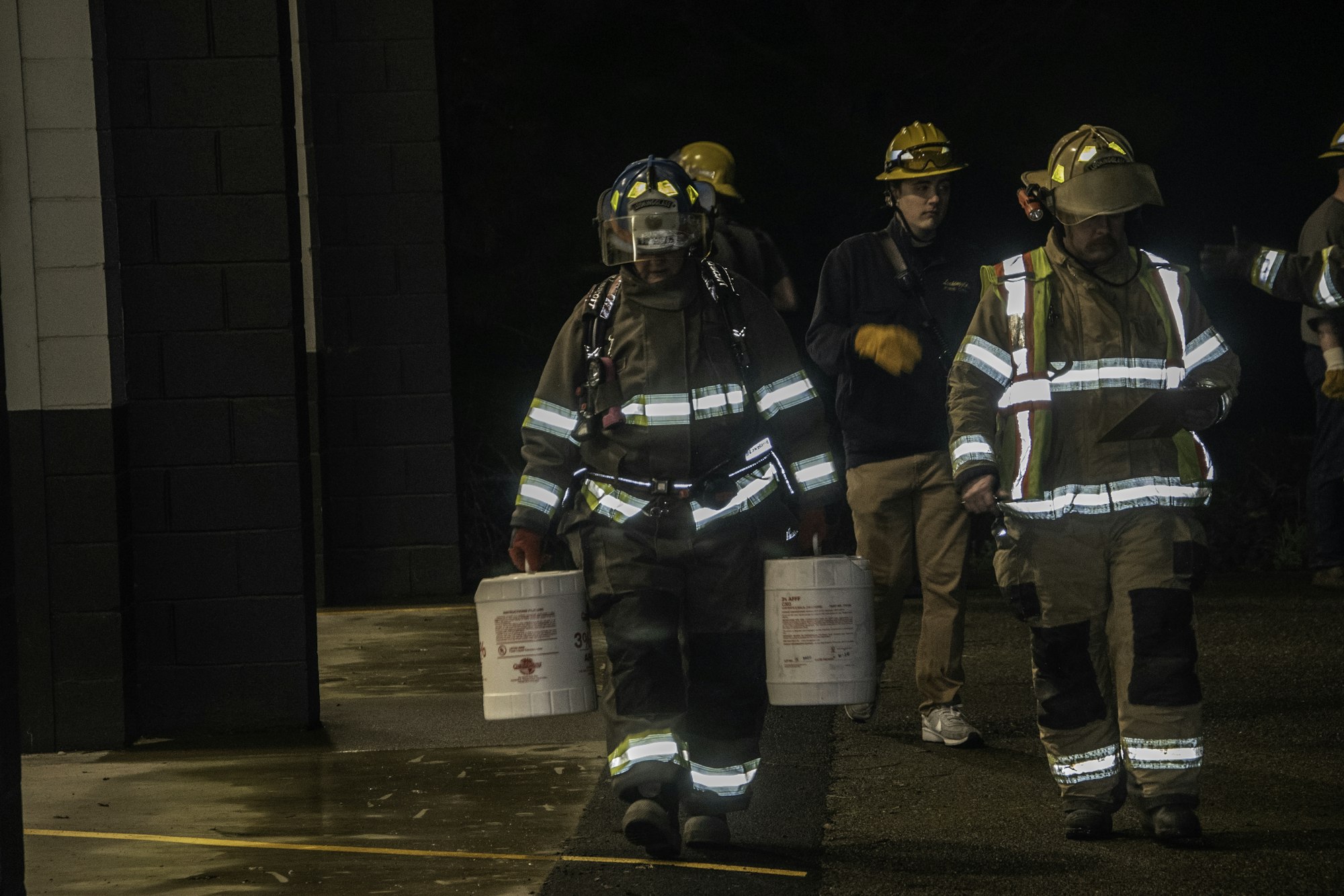 Firefighters in gear are walking outside, carrying containers and preparing for a task at night.