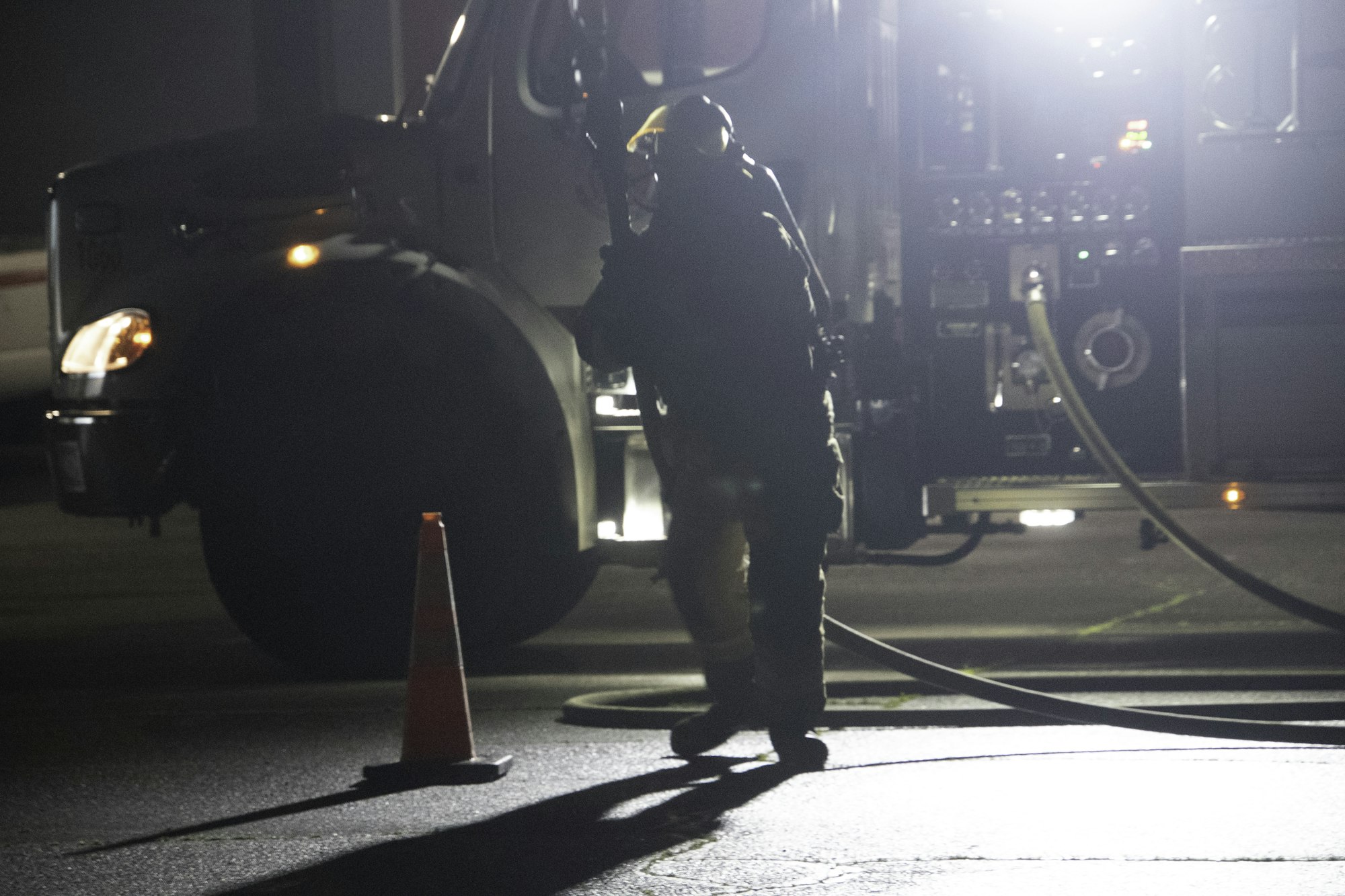 A figure in firefighter gear stands near a fire truck at night, with a traffic cone in the foreground and a hose on the ground.