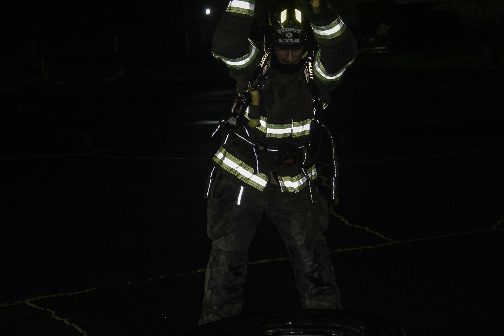 A firefighter in protective gear is training or working at night, lifting an object, possibly a tire, on a dark surface.