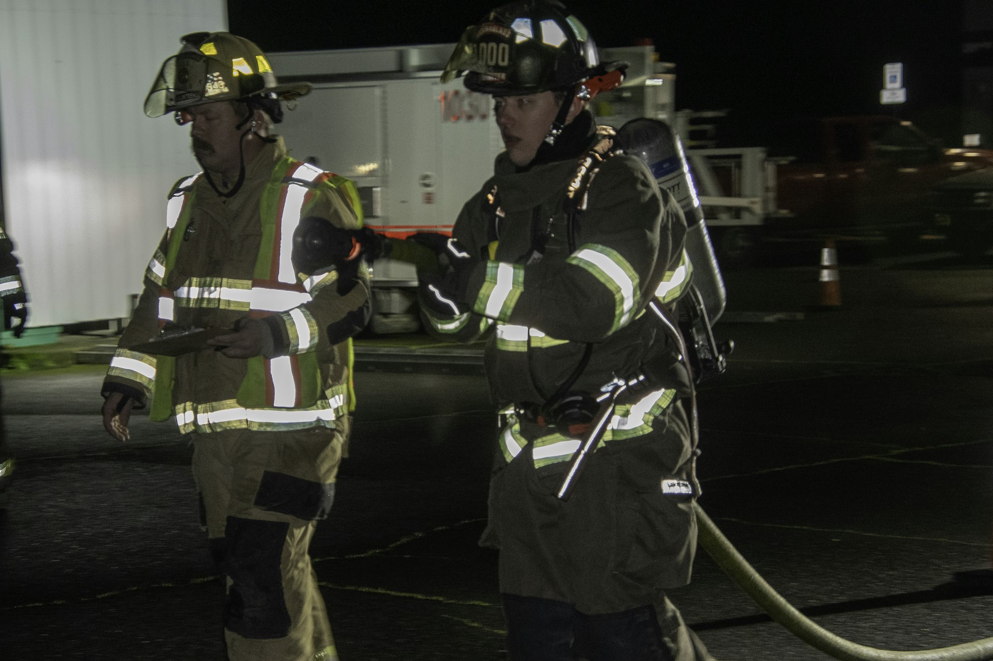 Two firefighters in gear walk together at night, one holding a clipboard and the other with an air tank and hose.