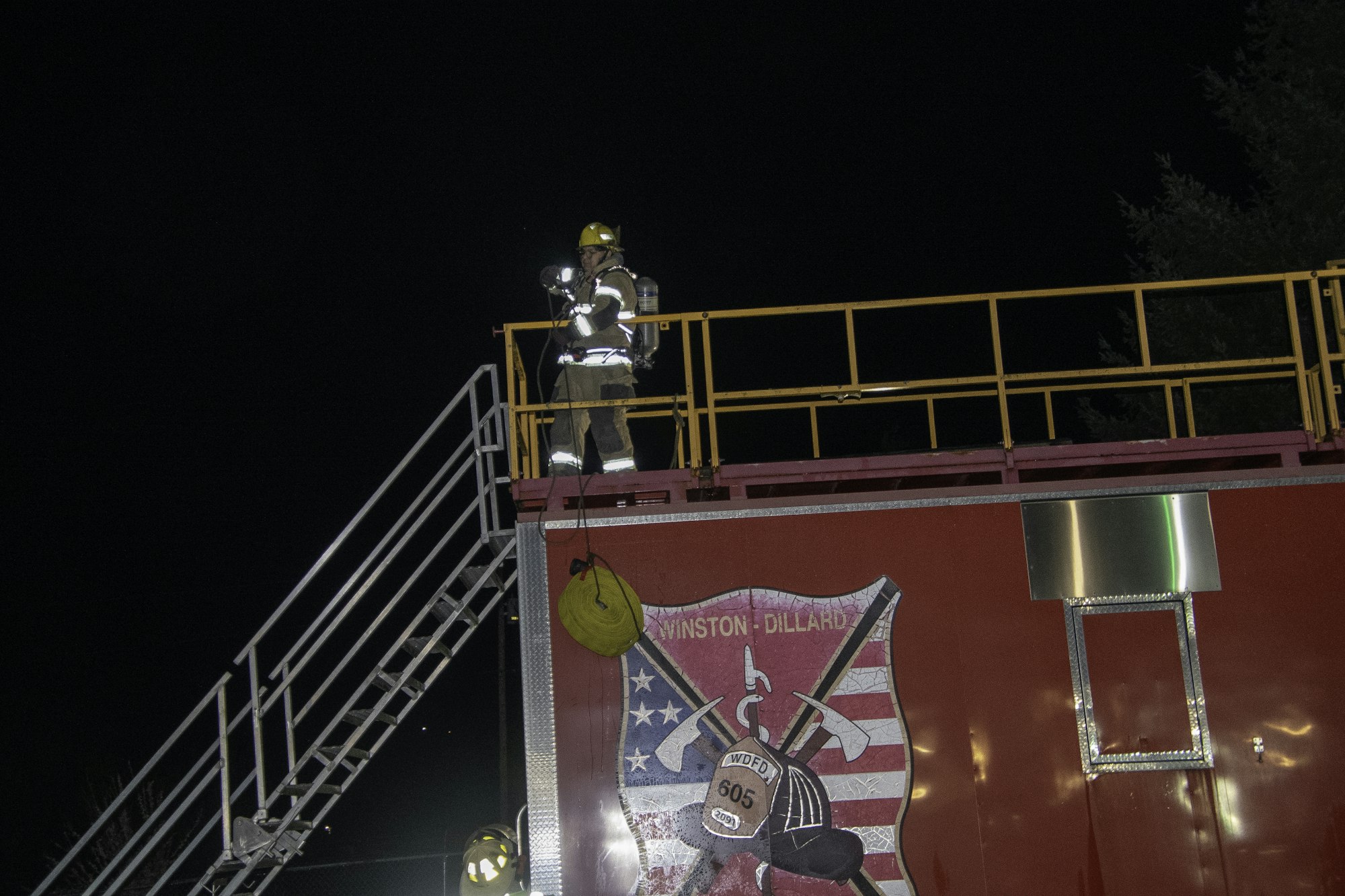 A firefighter in gear stands on top of a structure at night, with a ladder and equipment visible. A logo is on the side.