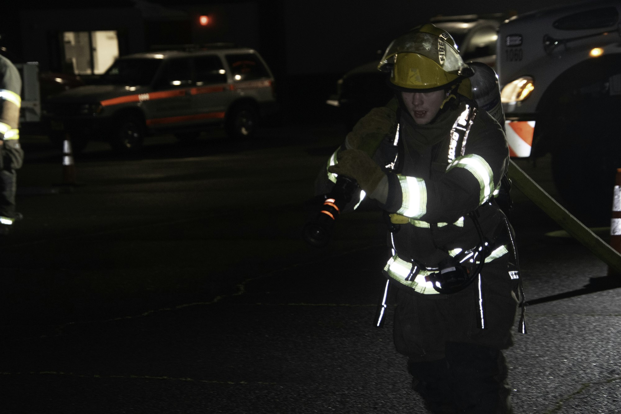 A firefighter in a dark environment, wearing gear and holding a flashlight, is preparing for action. Emergency vehicles are in the background.