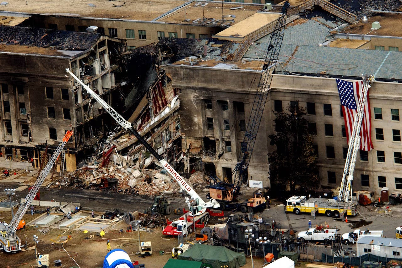 Damaged building with cranes, debris, and a large American flag hanging. Emergency response vehicles and equipment on site.