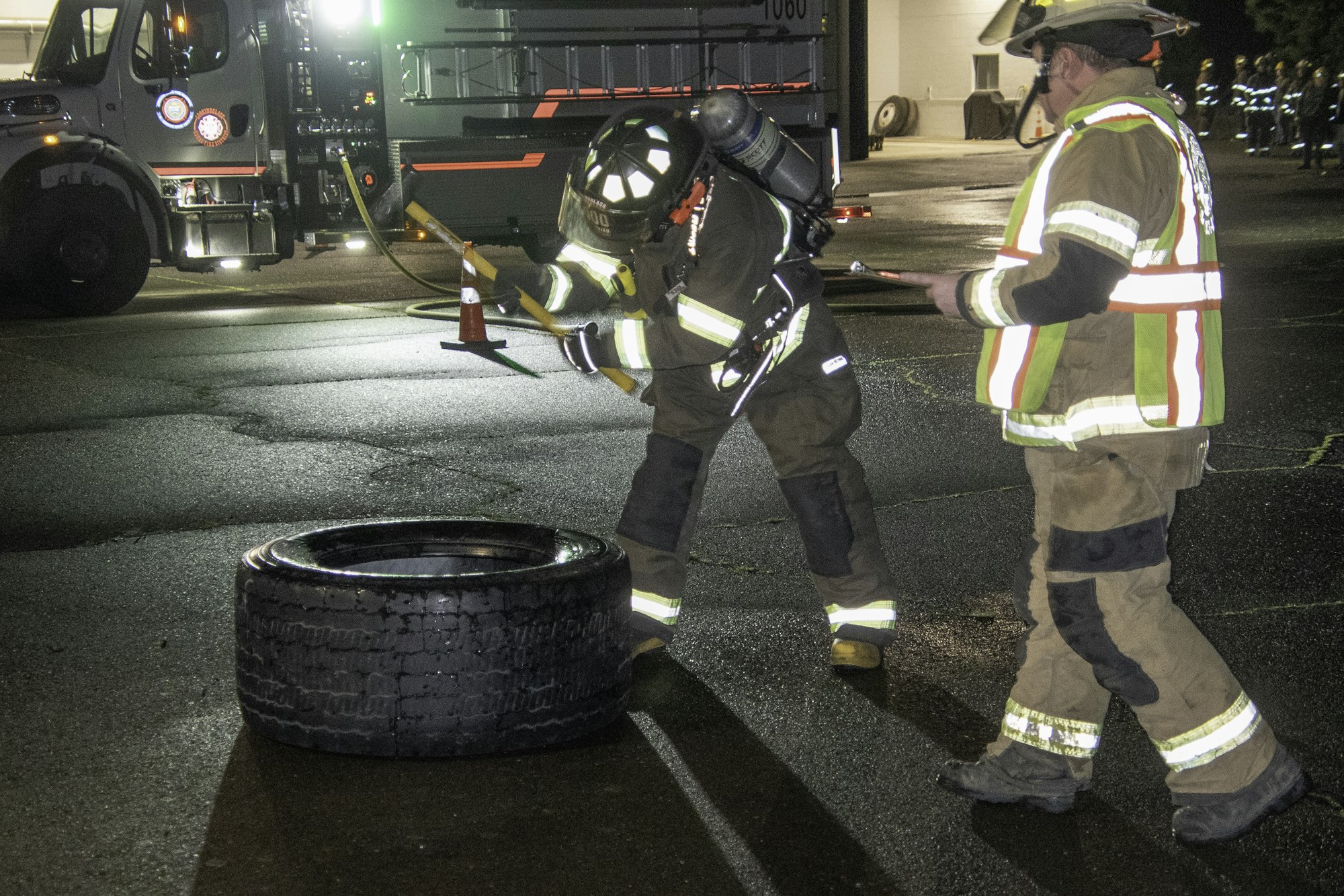 Firefighters in gear examine a tire on the ground at night, with a fire truck and other personnel visible in the background.
