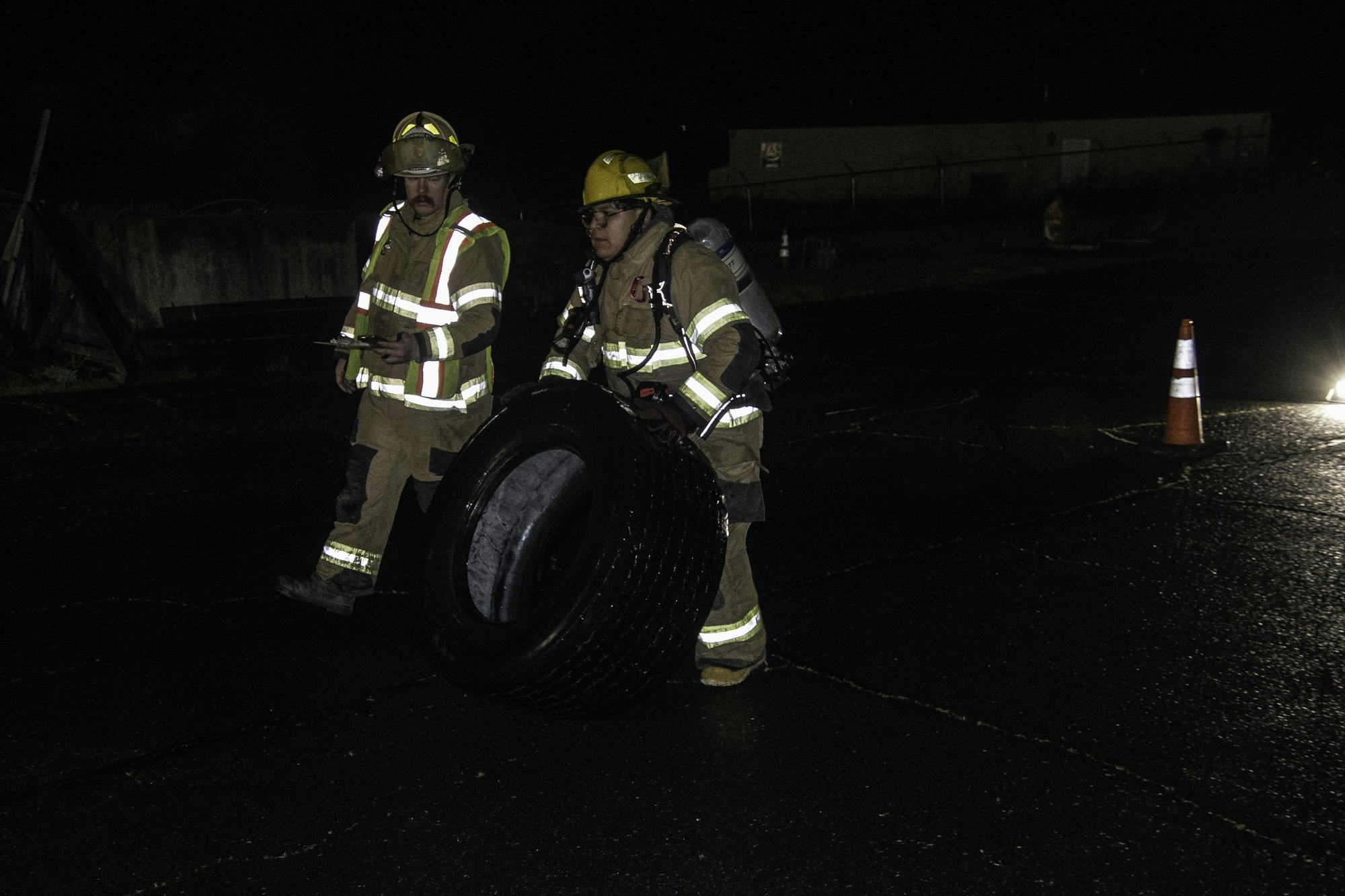 Two firefighters in gear, one holding a clipboard and the other carrying a large tire at night, with traffic cones nearby.