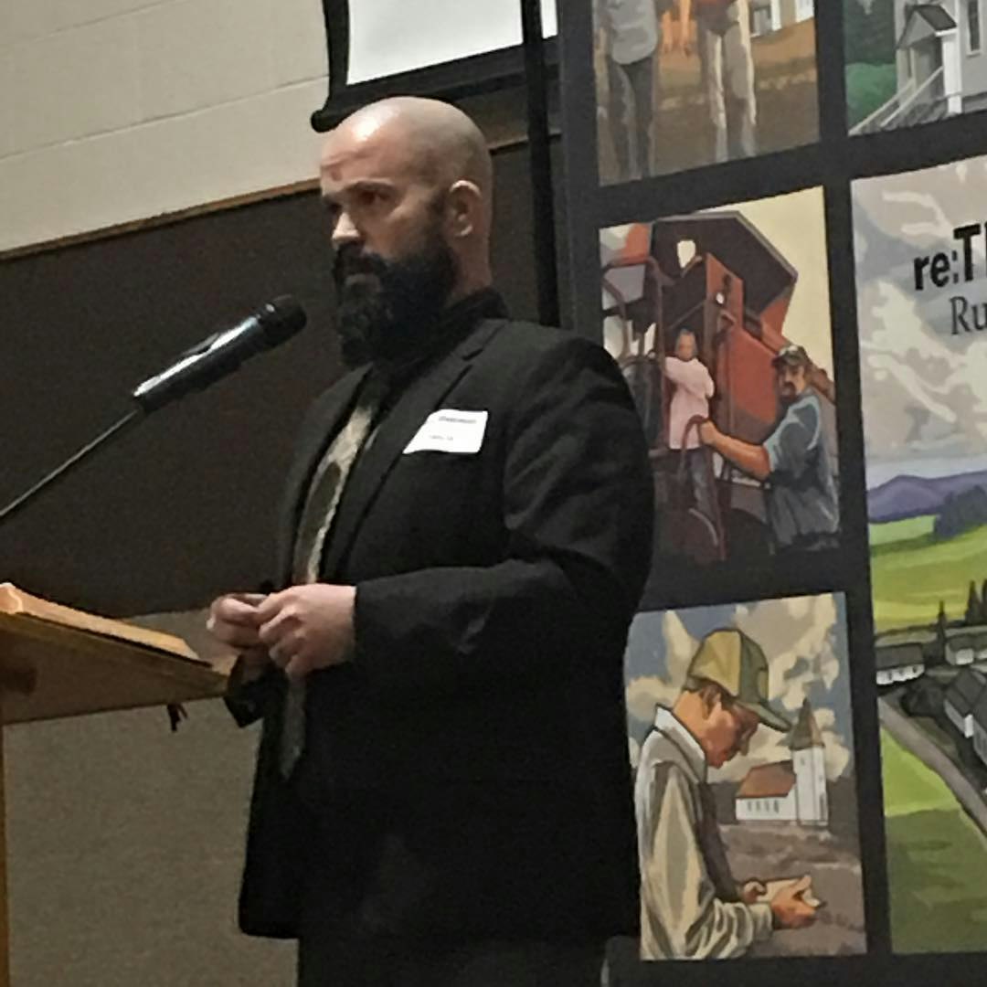 A bearded man stands at a podium with a microphone; a banner with illustrations in the background.