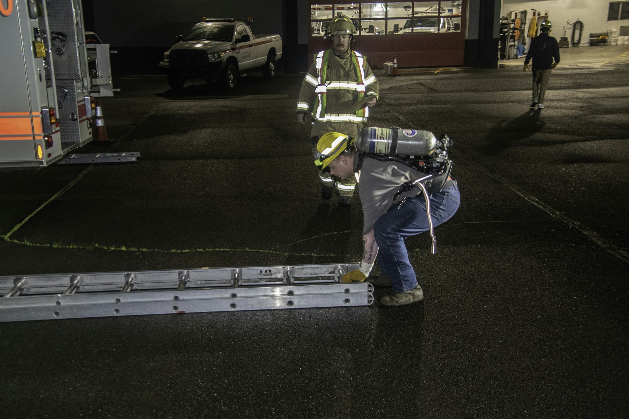 Two firefighters, one adjusting a ladder and another observing, with fire trucks in the background at night. Safety gear evident.
