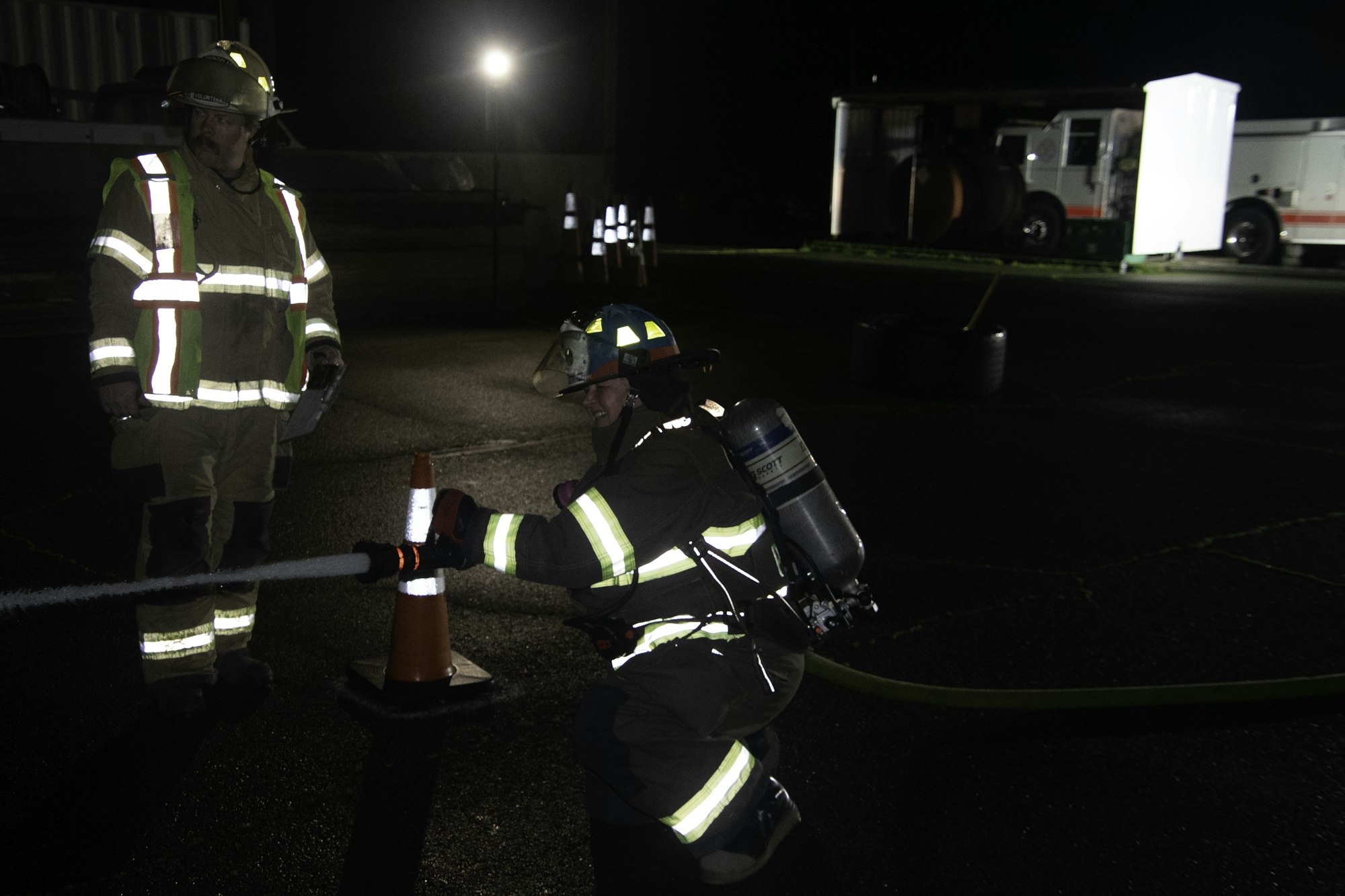 A firefighter in gear operates equipment near a traffic cone at night, while another observes nearby, likely during training.