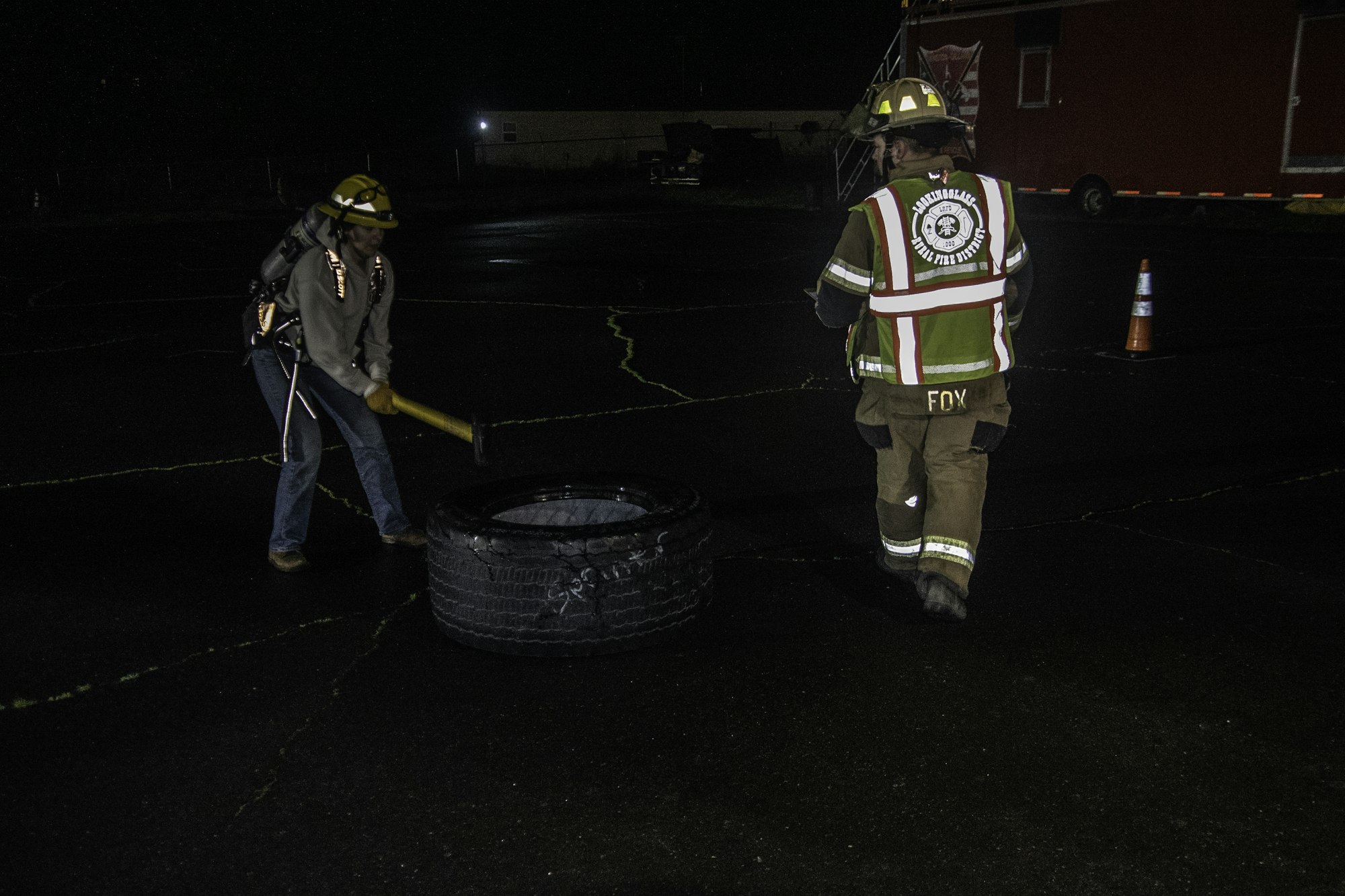 Two people are working at night, one swinging a hammer at a tire while the other observes, wearing fire gear.