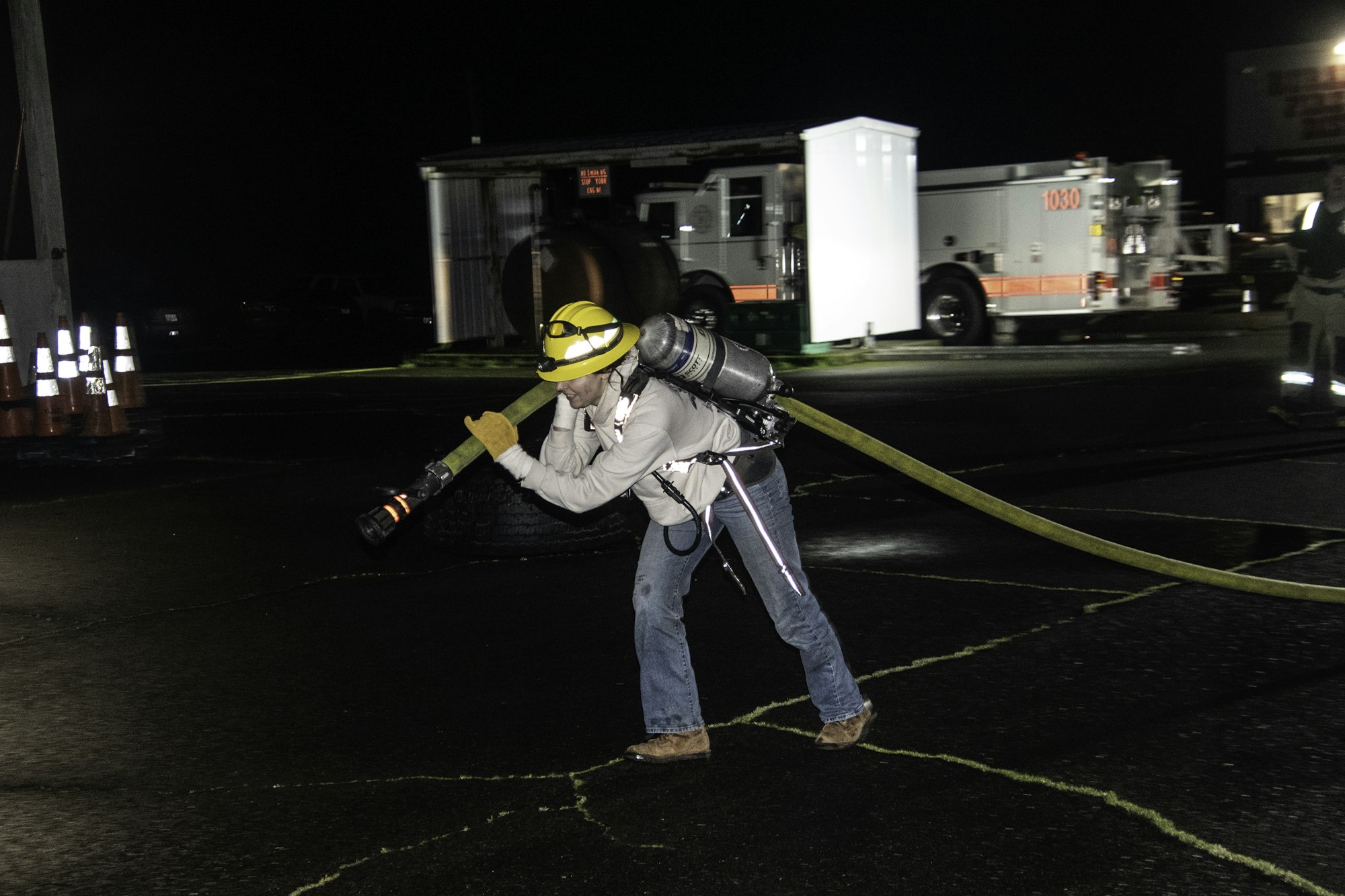 A firefighter in gear is dragging a hose at night, with emergency vehicles in the background and safety cones near the scene.