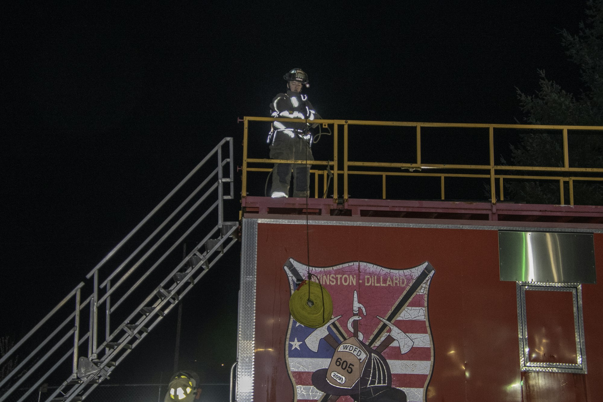 A firefighter stands on a platform at night, overlooking a fire truck with a logo and orange rescue rope hanging down.