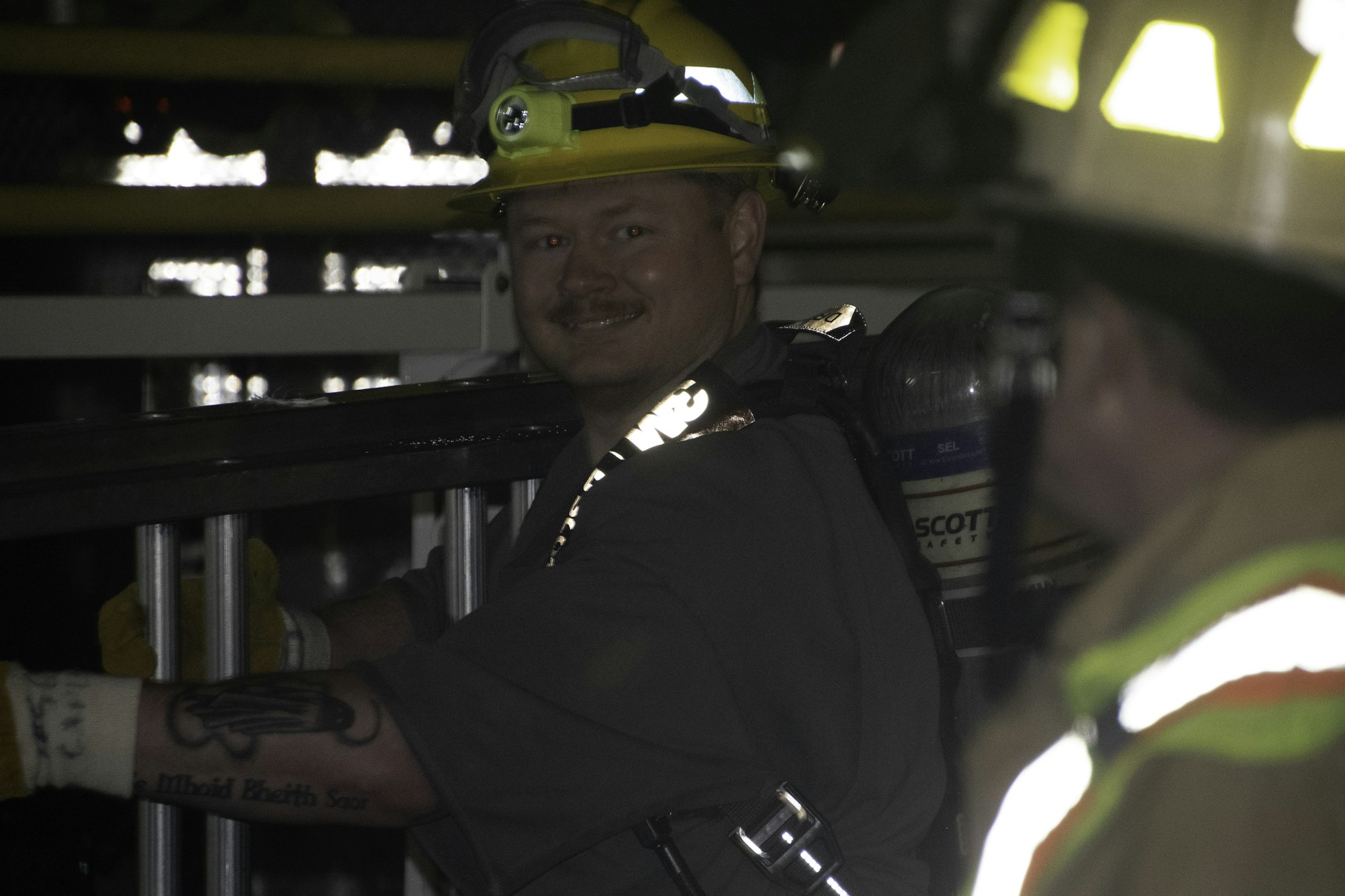 A firefighter wearing a helmet and safety gear smiles while handling equipment, with another person in the background.