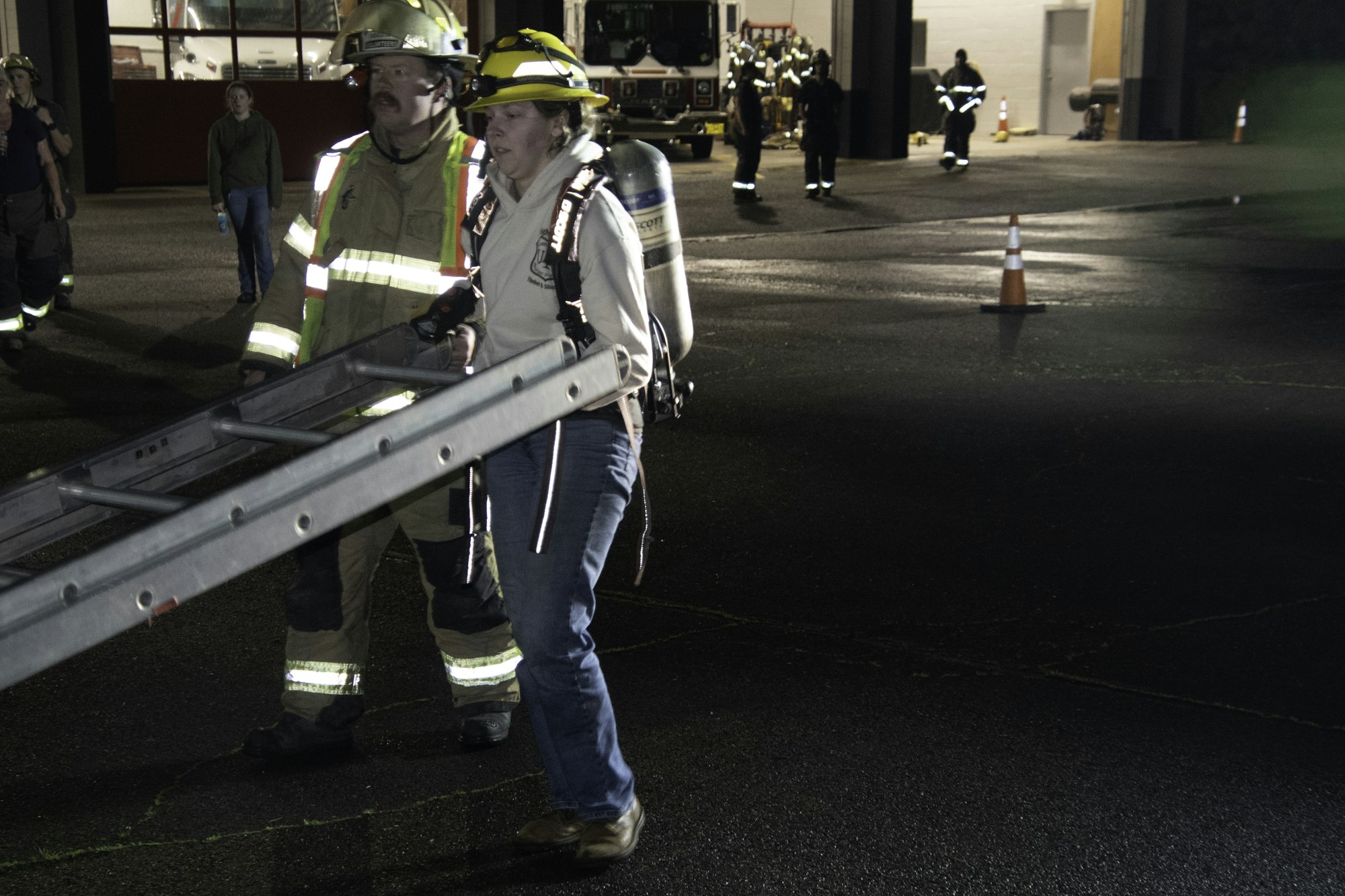 Firefighters and a volunteer are training, handling a ladder, in a well-lit area with emergency vehicles in the background.