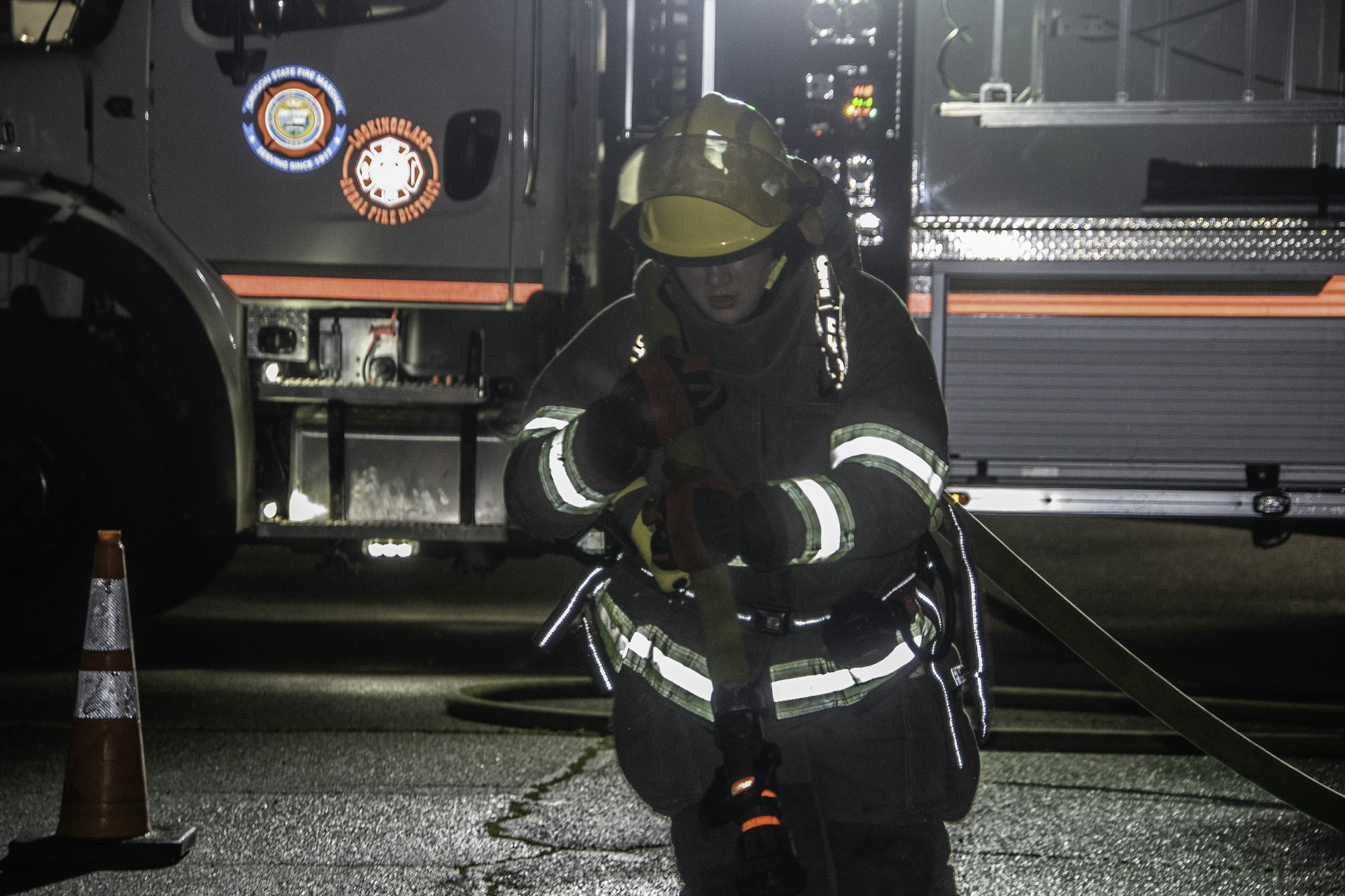 A firefighter in gear prepares a hose at night, with a fire truck illuminated in the background. Safety cones are present.