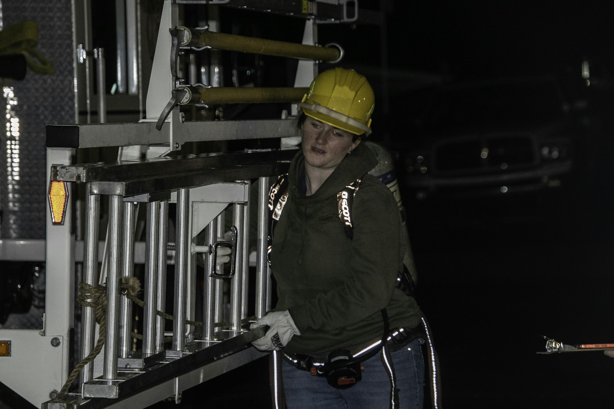 A person in a hard hat is handling a ladder near a fire truck at night, wearing safety gear and gloves.