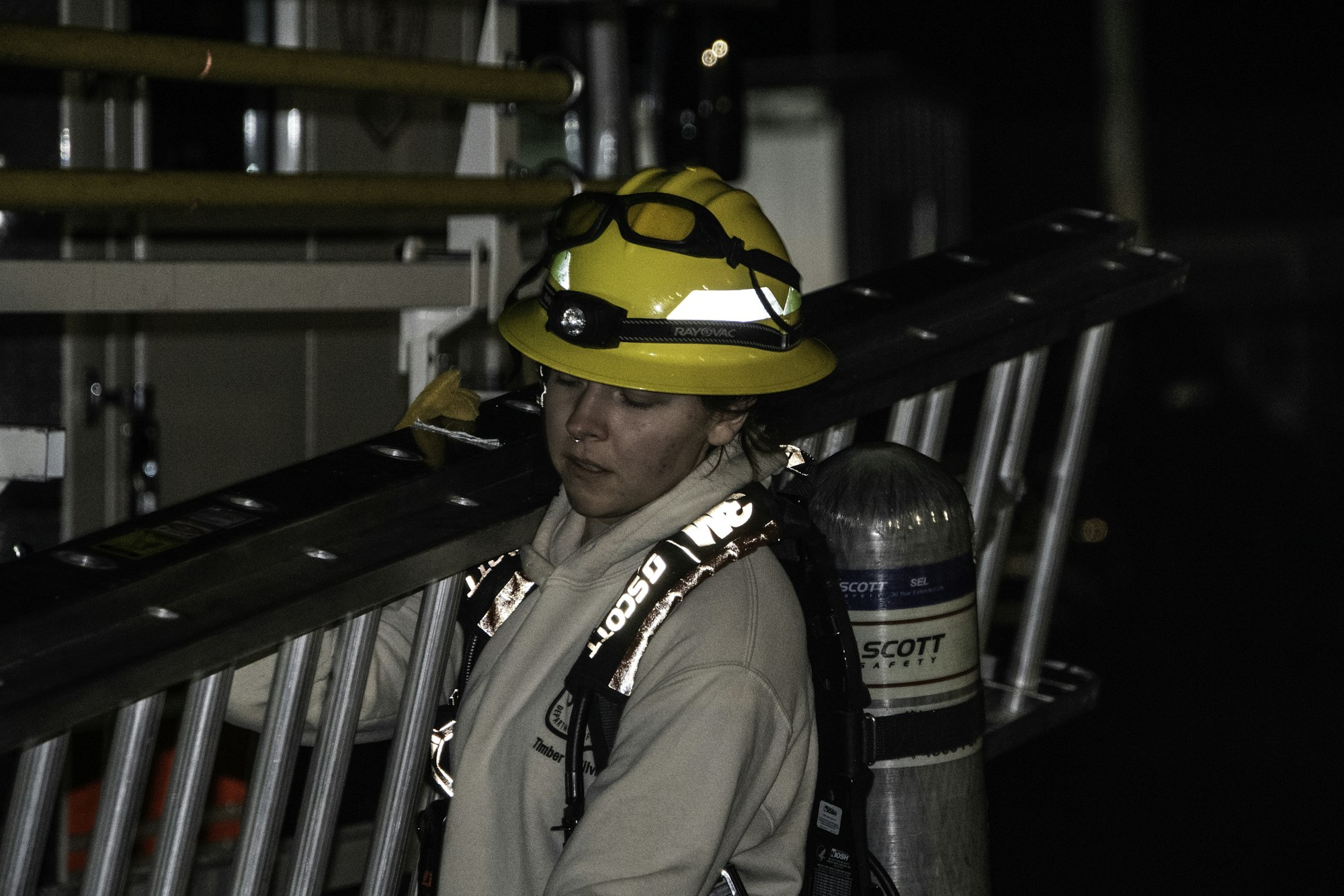 A firefighter in a yellow helmet carries a ladder at night, wearing safety gear and a harness.