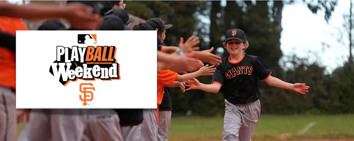 A kid in a Giants uniform is celebrating during a "Play Ball Weekend" event, high-fiving teammates on the field.