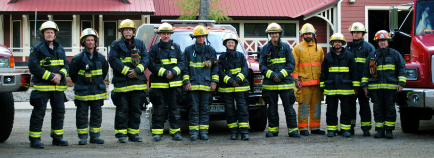 A group of firefighters in gear standing in front of fire trucks.