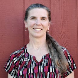A smiling woman with long hair, wearing earrings and a patterned blouse, standing against a red wall.