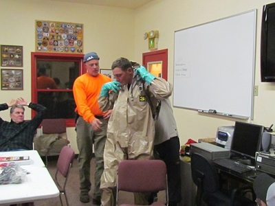 Two people assisting another person put on a hazmat suit indoors, with observers in the background.