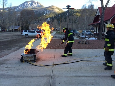 A firefighter is training, aiming a hose at a controlled blaze on a grill, another watches; mountains in the background.