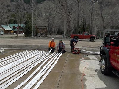 Three individuals working on what appears to be metal poles or beams next to a pickup truck in an outdoor setting with trees.