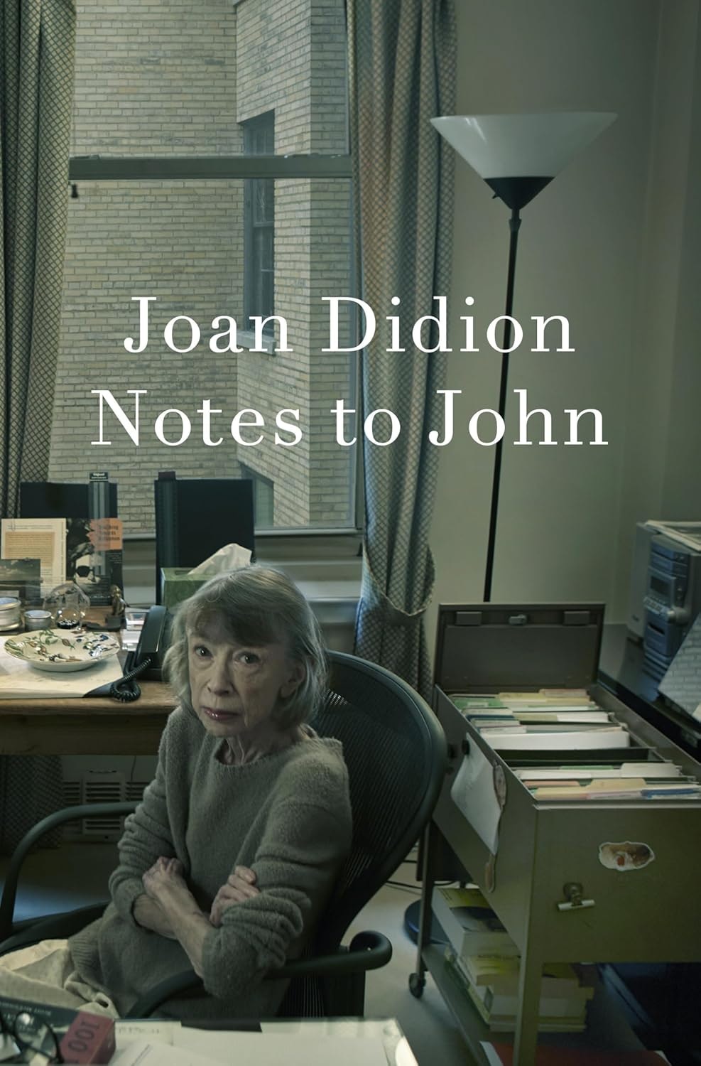 A woman sits at a desk in an office with the text "Joan Didion Notes to John" overlaid.