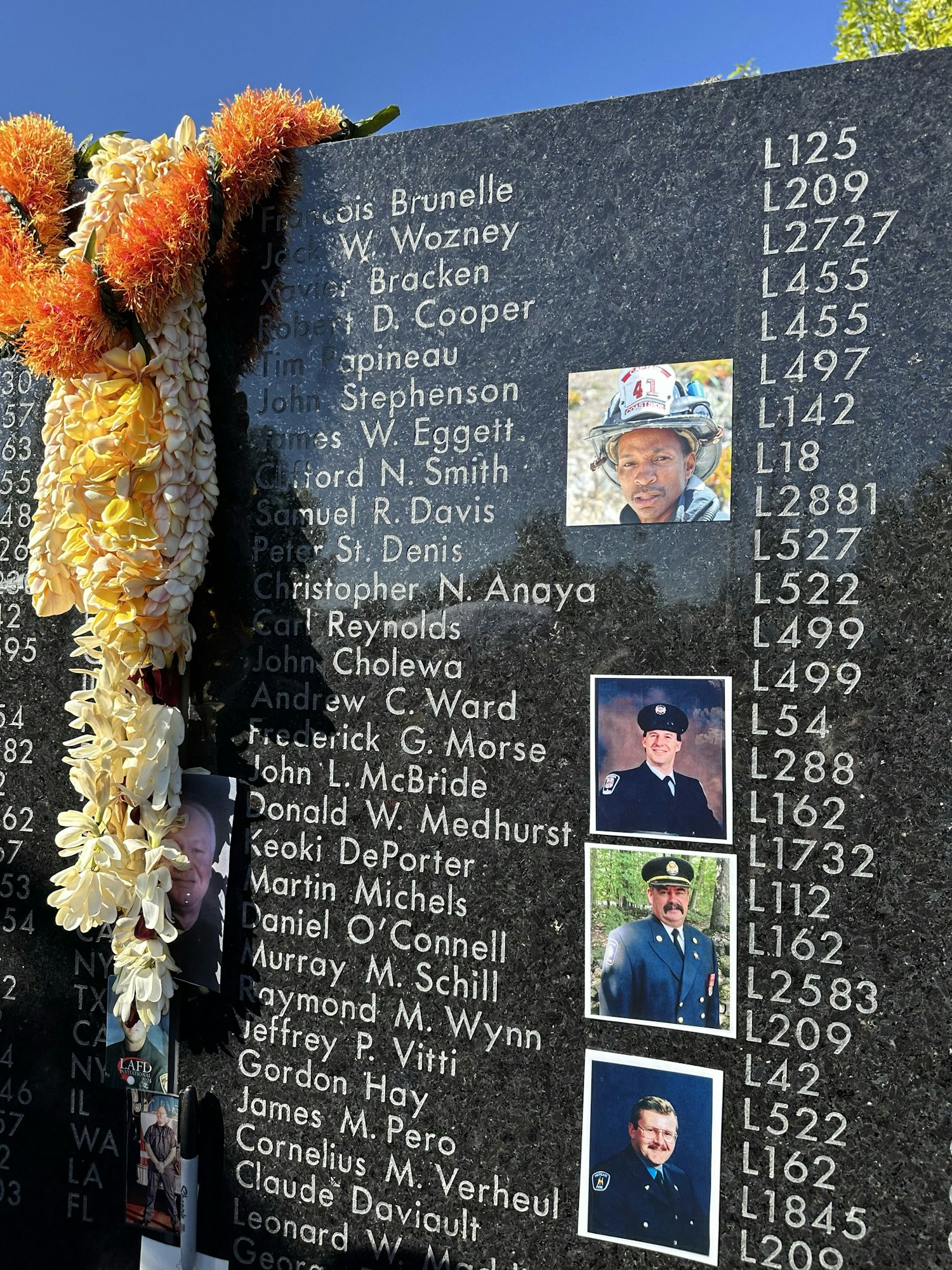 A memorial wall with names, codes, floral lei, and photos of uniformed individuals, suggesting tribute to fallen heroes or significant figures.