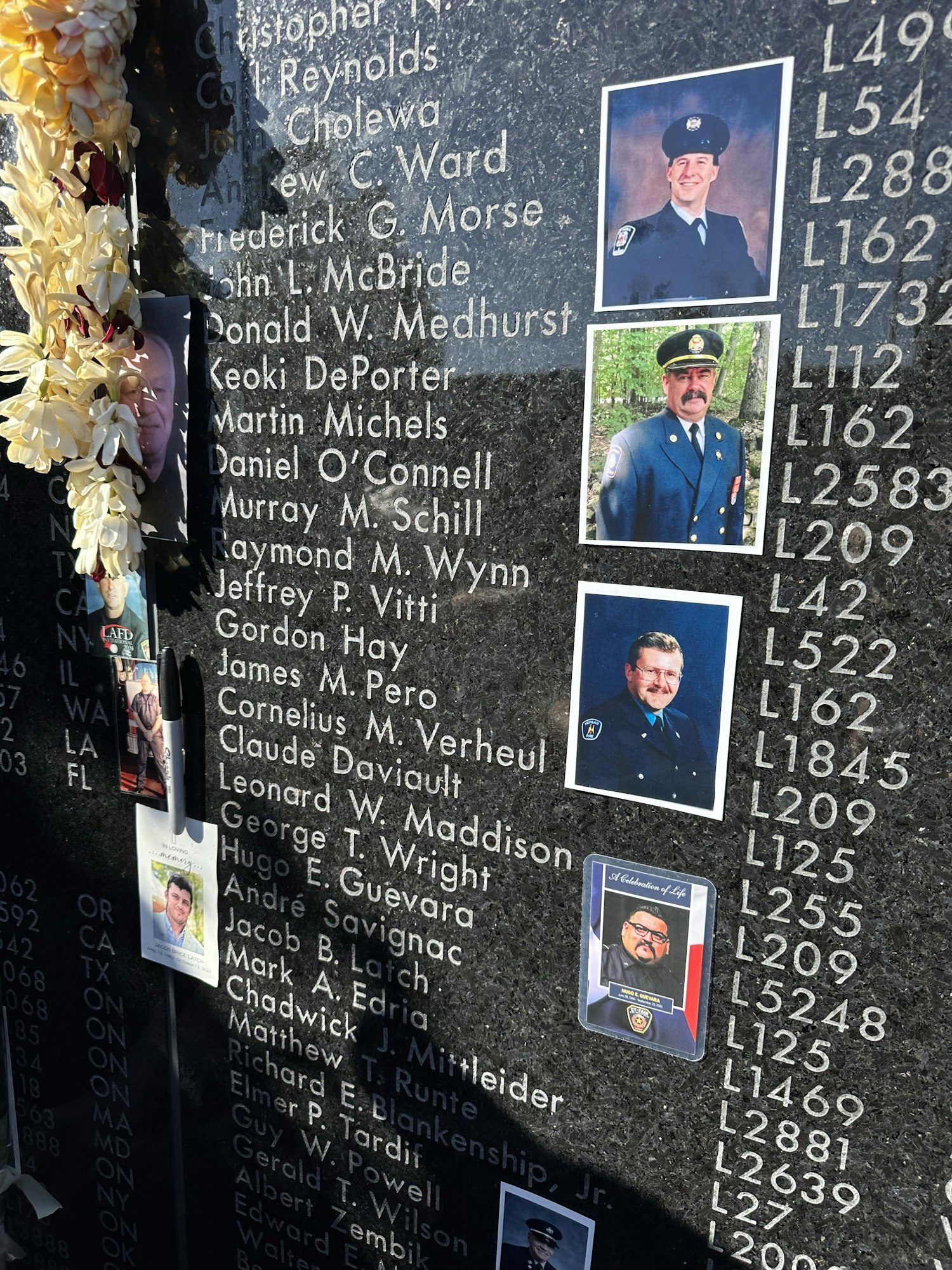 A memorial wall with names and photos, likely honoring deceased individuals, adorned with flower lei and personal mementos.